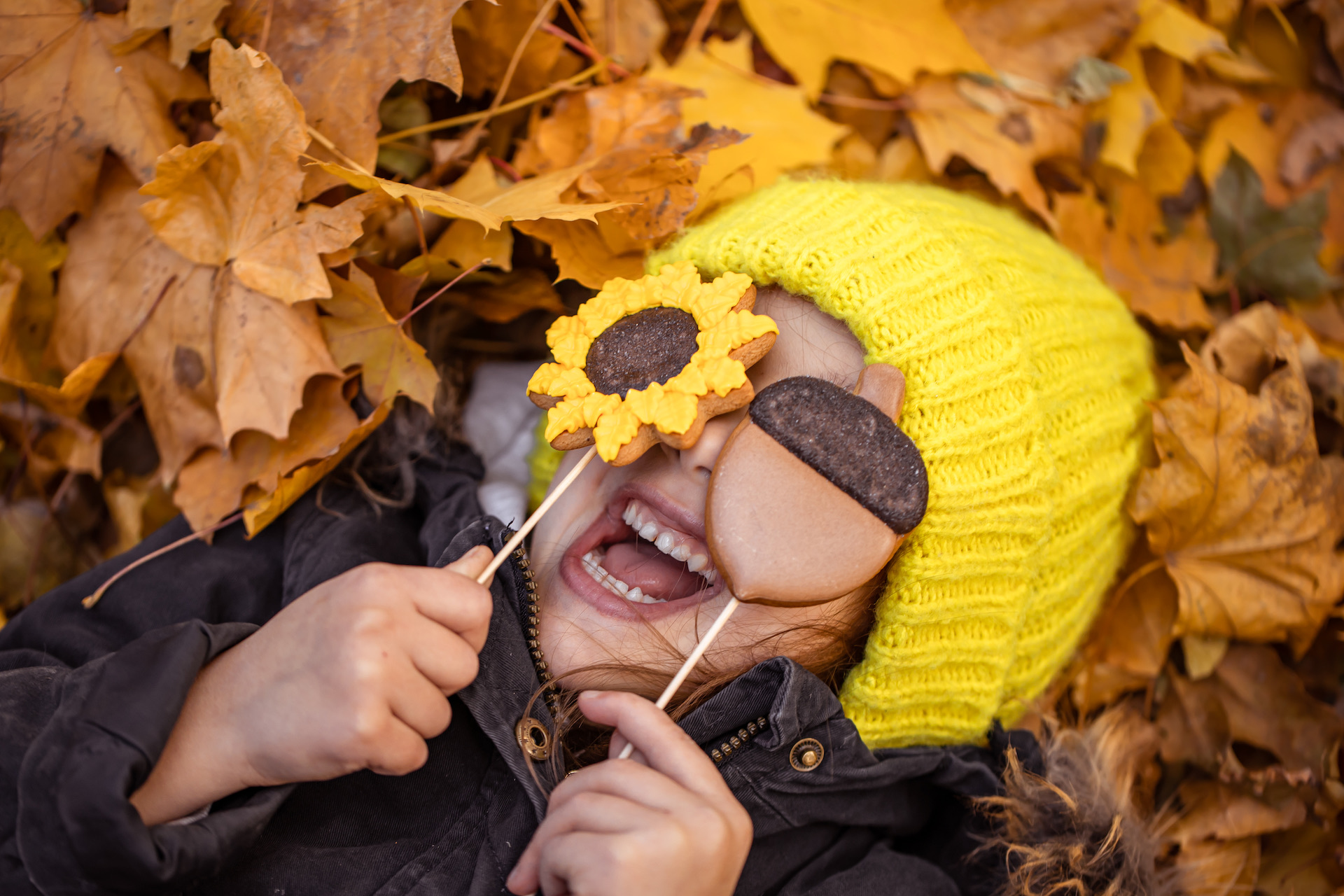 A little funny girl in a yellow hat lies in the autumn foliage and holds gingerbread in her hands.